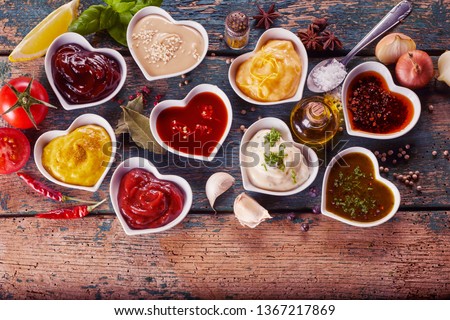 Assortment of marinades, sauces and dressings in individual heart shaped bowls surrounded by scattered spices, herbs and condiments on rustic wood
