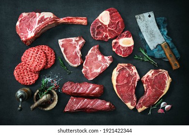 Assortment Of Raw Cuts Of Meat, Dry Aged Beef Steaks And Hamburger Patties For Grilling With Seasoning And Utensils On Dark Rustic Board