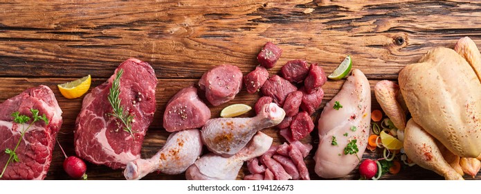 Assortment of raw meat on wooden table with vegetables, greens and sprinkled with spices, viewed from above with copy space