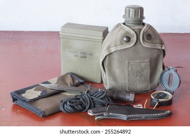 Assortment Of Survival Hiking Gear Including First Aid Kit, Canteen, Knife, Parachute Cord, Compass And Map Holder On Old Wood Table