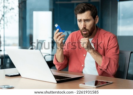 Asthma and respiratory problems in office worker, mature man coughing and using medicine in inhaler to ease breathing at workplace, mature businessman inside office with laptop.