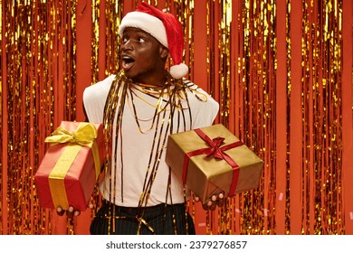 astonished african american man in santa cap holding gifts and looking away near golden shiny tinsel - Powered by Shutterstock