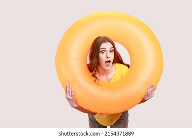 Astonished Amazed Female Young Age With Brown Hair Holding Orange Rubber Ring, Looking At Camera With Shocked Expression And Open Mouth. Indoor Studio Shot Isolated On Gray Background.