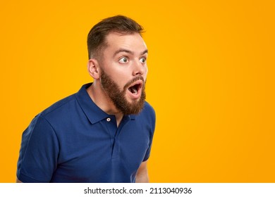 Astonished bearded man in blue t shirt looking away with opened mouth against yellow background