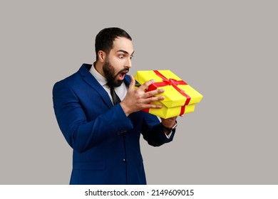 Astonished bearded man unpacking present, looking inside box with shoked and scared expression, birthday gist, wearing official style suit. Indoor studio shot isolated on gray background.