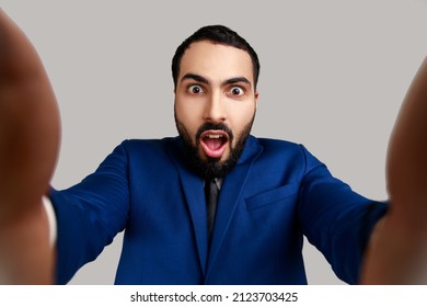 Astonished bearded young adult man taking selfie, looking at camera with shocked expression and open mouth POV, wearing official style suit. Indoor studio shot isolated on gray background.
