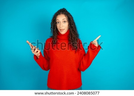 Astonished Beautiful girl with curly hair wearing red knitted sweater holding her telephone and pointing with finger aside at empty copy space