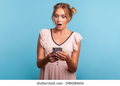 Astonished beautiful young adult woman in summer dress, using smartphone and watching shocking video, reading message with amazed expression. Indoor studio shot isolated on blue background.