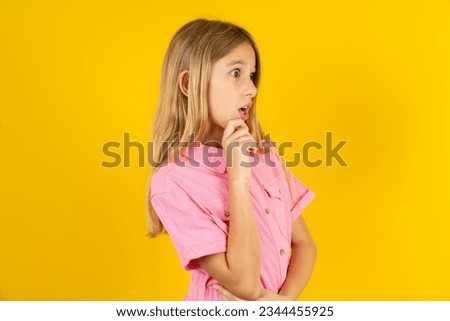 Astonished Caucasian kid girl wearing pink dress over yellow background looks aside surprisingly with opened mouth.