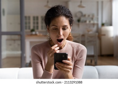 Astonished Hispanic Young 30s Woman Sit On Couch At Home Looks At Smart Phone Screen Open Mouth Read Unbelievable News, Cant Believe In Luck, Got Fantastic Offer, Low Price Huge Discount, Sale Concept