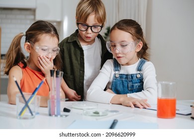 Astonished Little Kids Doing Home Science Project, Looking At The Glass Dish In Amasment. All Wearing Glasses. Chemical Glassware And Colored Liquids On The Table.