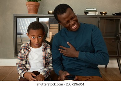 Astonished little preschool boy with brown skin looking at camera with jaw dropping after hearing adult joke from his father sitting next to him on floor laughing out loud, holding phone in hands