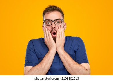 Astonished male in glasses and blue t shirt expressing surprise after hearing shocking news while looking at camera with opened mouth on yellow background