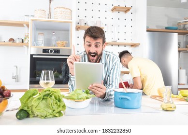 Astonished man using digital tablet while friend is busy preparing food in the kitchen