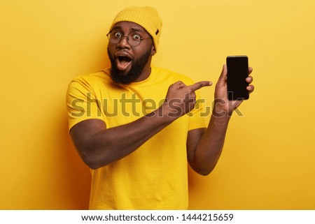 Astonished plump dark skinned man holds mobile phone with copy empty screen, points at device, surprised with high quality gadget and its functions, wears yellow hat and t shirt. Selective focus