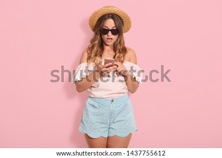 Astonished pretty woman wears blouse with bared shoulders, short, straw hat and sunglasses posing with open mouth, holds her cell phone with surprised facial expression, recives messages from friend.