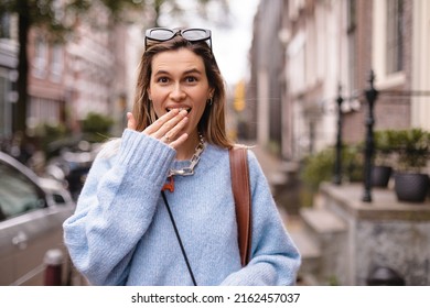 Astonished and shocked young woman in casual clothes covering her mouth with hand, looking at camera with amazed smiling facial expression walking on the street, urban. Human positive emotions concept