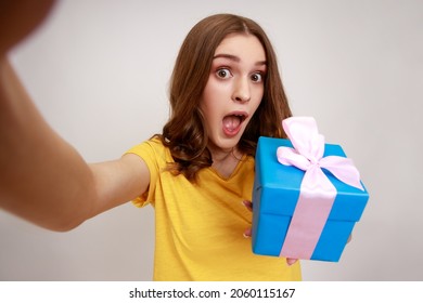 Astonished Teenager Girl In Yellow Casual Style T-shirt With Widely Opened Mouth Looking At Camera POV, Point Of View Of Photo, Holding Present Box. Indoor Studio Shot Isolated On Gray Background.