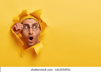 Astonished unshaven man gazes through hole in yellow paper, wears round spectacles, opens mouth with surprisement, copy space to insert text or slogan. Effect of torn paper. Discount and sale