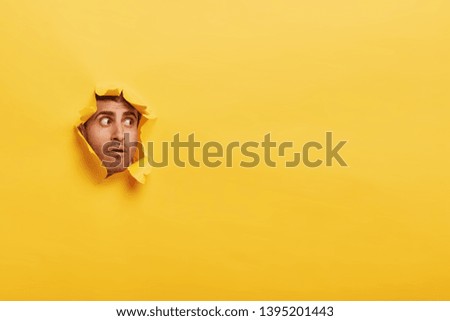 Astonished unshaven man has stupefied expression, peeks through torn wall paper, has widely opened eyes, stares through hole in yellow background. Human facial expressions and emotions concept