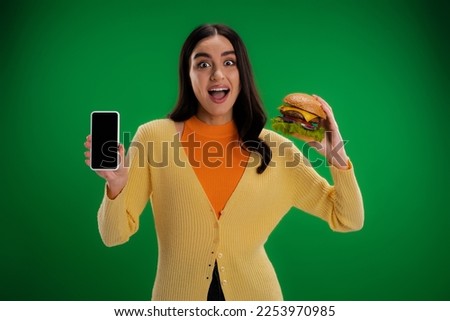 astonished woman holding tasty burger and mobile phone with blank screen isolated on green