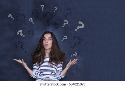 Astonished young girl standing against blackboard wall with question mark sketches on it. Concept of questions without answers.
