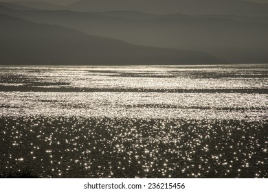 An astonishing artistic view of black water glimmering with silvery light with gray mountains in the horizon.