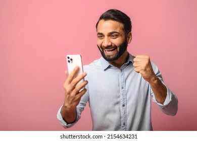 Astonishment Indian Man Using A Smartphone Isolated On Pink Background, Hispanic Guy Looks At The Screen With Mouth Opened Holding Phone, Excited With Good Deal Or Unexpected News