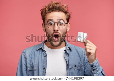 Astonsihed male wears spectacles, opens mouth with great surprisment as finds condom in bedroom, doesn`t know who used it. Shocked adult holds rubber condom, isolated over pink studio background