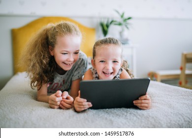 Astrakhan, Russia - July 18, 2019: Two beautiful little sisters lying in the bed and look at the screen of a tablet, smart kids using smart technology