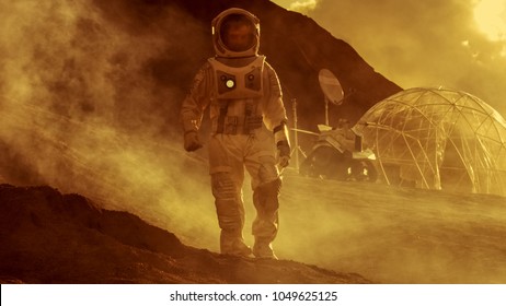 Astronaut On Mars Walking On The Exploring Expedition. In The Background His Base/ Research Station. First Manned Mission To Mars, Technological Advance Brings Space Exploration, Colonization.