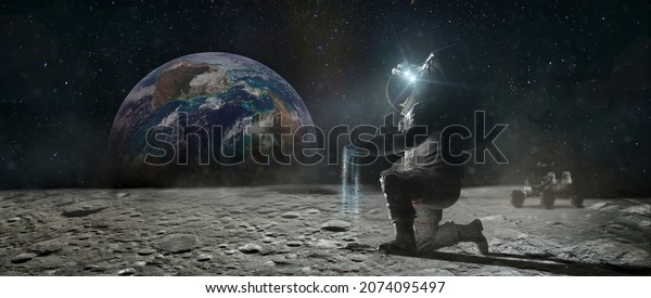 Astronaut sits on his knee\
on the lunar surface and pours blue earth\'s soil through his\
fingers. Planet Earth rise at the horizon. Elements of this image\
furnished by NASA.