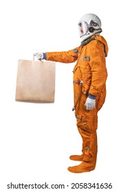 Astronaut Wearing Orange Space Suit And Space Helmet Holding In Hand Brown Clear Empty Blank Craft Paper Bag For Takeaway Isolated On A White Background. Delivery Or Takeaway Concept. Side View.