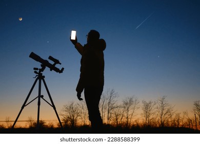 Astronomer looking at the starry skies with planets, falling stars and Moon eclipse with a telescope while using modern smartphone. - Powered by Shutterstock