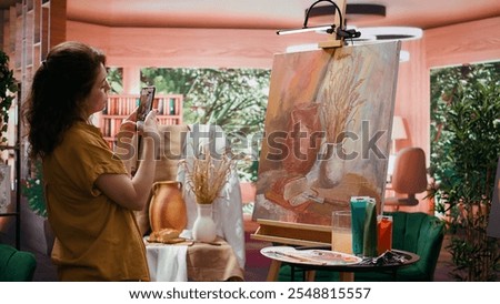 In atelier, painter is promoting her rustic masterpiece painting with the smartphone, self advertising content on social media. Filming colorful canvas with fine art piece. Online presence. Camera B.