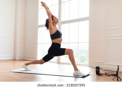 Atheltic Young Woman Doing Forward Lunges Exercise With Arms Overhead Watching Online Workout Video Tutorial Using Cell Phone, Standing In 1 Warrior Pose On Yoga Mat. Domestic Female Fitness Concept