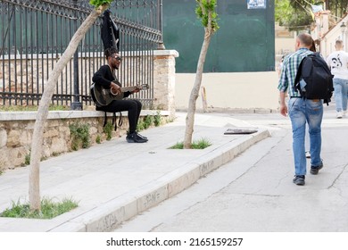 Athens, Greece, 04.30.2022. A Tourist Walks Through The City.
Nearby, A Black Street Musician Sits Along The Sidewalk And Plays The Guitar.