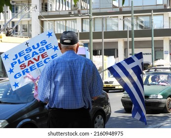 Athens, Greece-May 18, 2022: Close up of elderly man holding up a sign;"Jesus please save Greece" and a Greece flag on a busy street in Athens.