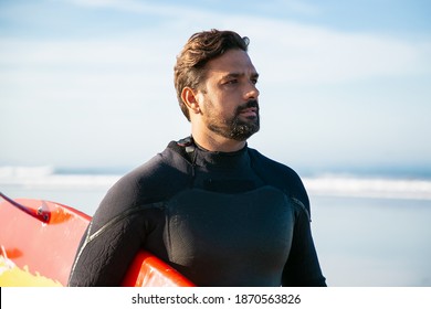 Athlete in black wetsuit holding surfboard and looking away. Portrait of serious attractive brunette surfer standing at coast near sea. Lifestyle, vacation and summer activity concept - Powered by Shutterstock