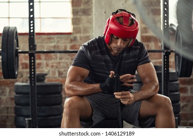 Athlete Boxer In Sportswear Tying Bandage On Hand And Wearing Red Boxing Helmet. Mature Boxer With Head Gear Wearing Hand Gloves. African Man Preparing For Boxing Practice At Gym. 