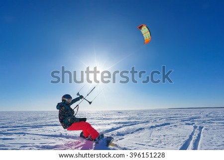 Athlete in bright clothes riding in the snow on a snowboard and kite controls. Deep blue sky and sun in the background
