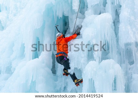 Athlete climbing cliff covered with ice, using ice axes, and piercing front crampons right onto the ice