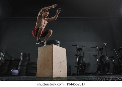 Athlete doing box jump exercise at the crossfit gym. Man jumping onto the box on gray wall background. Legs training. Copy space