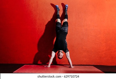 Athlete doing push ups on his hands while standing upside down near red wall. Full body length portrait