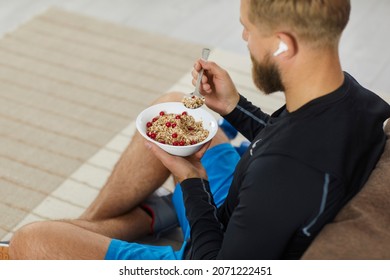 Athlete enjoying healthy meal rich in fiber, protein and vitamins. Fit young man sitting on floor in living room, relaxing after fitness workout, eating natural vegetarian granola, listening to music - Powered by Shutterstock