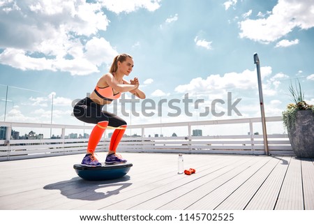 Athlete girl is enjoying work out with outfit on high balcony. She is doing squats on bosu platform while stretching resistance band under knees. Copy space in right side