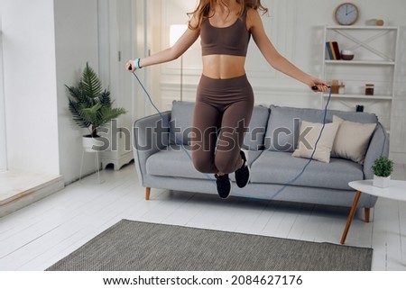 A athlete is jumping while holding her jump-rope. She is wearing dark sneakers. Her mat is gray. There is a gray sofa behind her. She is also wearing light-brown gym clothes.