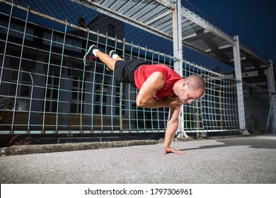 Athlete In A Red T-shirt Doing One Arm Handstand Push Ups At The Outdoor Gym.