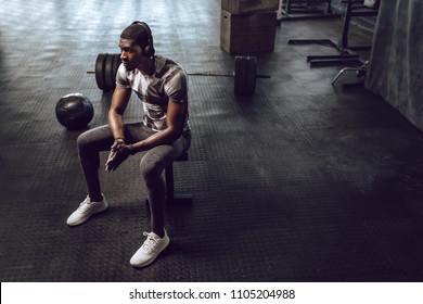 Athlete Relaxing After Workout Sitting On A Bench In The Gym. Man Listening To Music Using Wireless Headphones And Relaxing At The Gym.