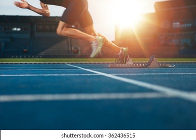 Athlete Running On Athletic Racetrack. Low Section Shot Of Male Runner Starting The Sprint From The Starting Line With Bright Sunlight.